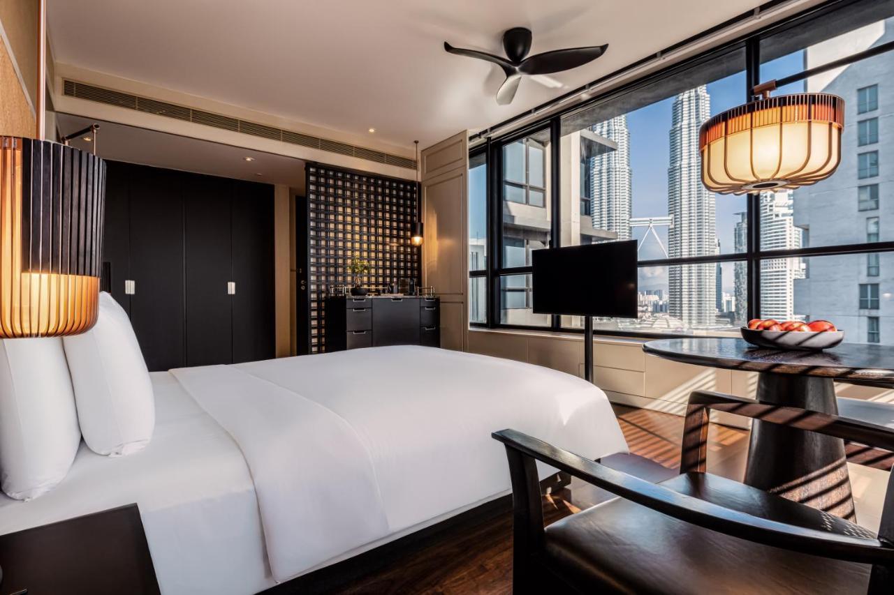 ザ ルーマ ホテル アンド レジデンシーズ クアラルンプール エクステリア 写真 The photo shows a modern hotel room featuring a large bed with white linens. There's a ceiling fan above and large windows letting in natural light, providing a view of a cityscape outside. To one side, there's a small seating area with a round table